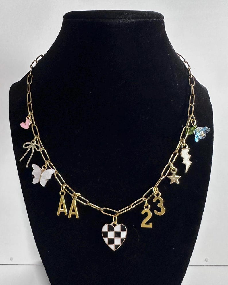 May include: A gold-toned chain necklace featuring various charms including a pink heart, a bow, a white butterfly, the letters AA, a black and white checkered heart, the numbers 23, a star, a lightning bolt, and a blue flower with a green leaf.
