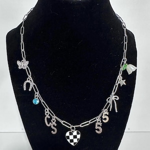 May include: A silver-toned chain necklace featuring various charms including a butterfly, a horseshoe, a blue crystal, the letters C and S, a black and white checkered heart, the number 5, a bow, a star, and a white flower with green leaves.