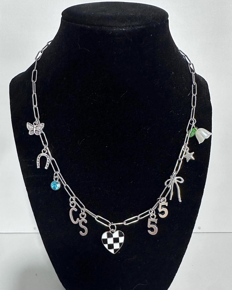 May include: A silver-toned chain necklace featuring various charms including a butterfly, a horseshoe, a blue crystal, the letters C and S, a black and white checkered heart, the number 5, a bow, a star, and a white flower with green leaves.