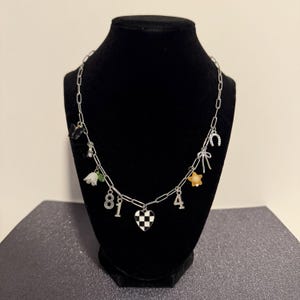 May include: A silver-toned chain necklace featuring multiple charms including a black butterfly, a white flower, the numbers 8, 1, and 4, a checkered heart, a yellow star, a silver bow, and a horseshoe. The charms are attached to a link chain.