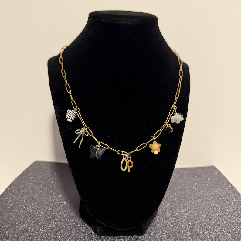 May include: A gold-toned chain necklace featuring multiple charms including a black and white checkered cube, a bow, a blue butterfly, the letters OP, a yellow star, a small black circle, and a white flower with a green leaf accent.