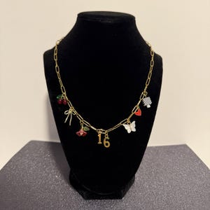 May include: A gold-toned chain necklace featuring multiple charms including a pair of red cherries, a bow, a red flower, the number 16, a white butterfly, a small red heart, and a black and white checkered square charm hanging from the links.