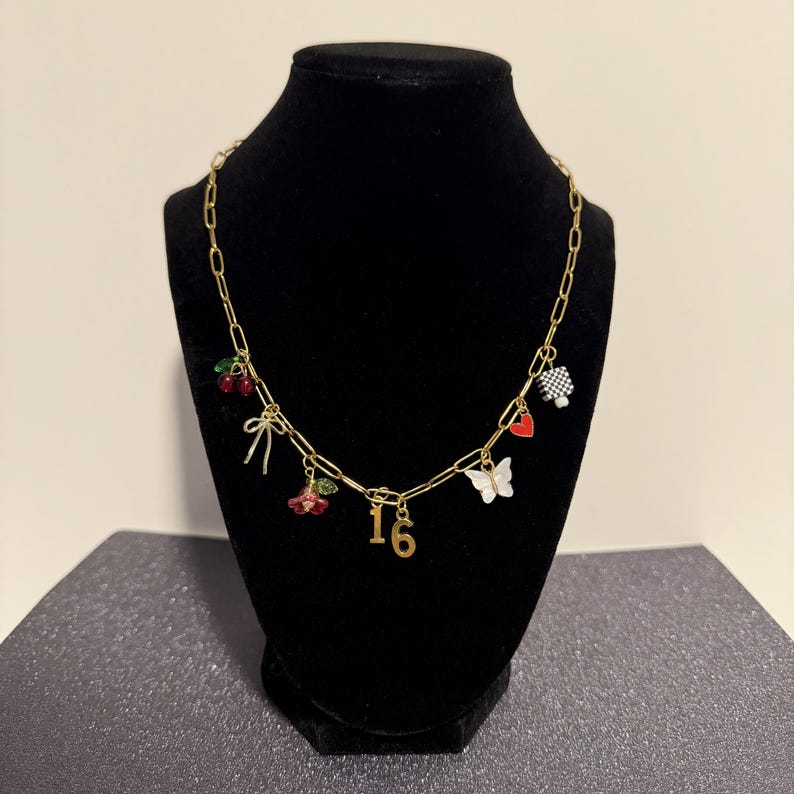 May include: A gold-toned chain necklace featuring multiple charms including a pair of red cherries, a bow, a red flower, the number 16, a white butterfly, a small red heart, and a black and white checkered square charm hanging from the links.