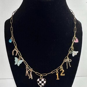 May include: A gold-toned chain necklace featuring various charms including a blue rhinestone, a horseshoe, a light blue butterfly, the letters KA, a black and white checkered heart, the number 12, a gold bow, a white flower, and a pink heart.