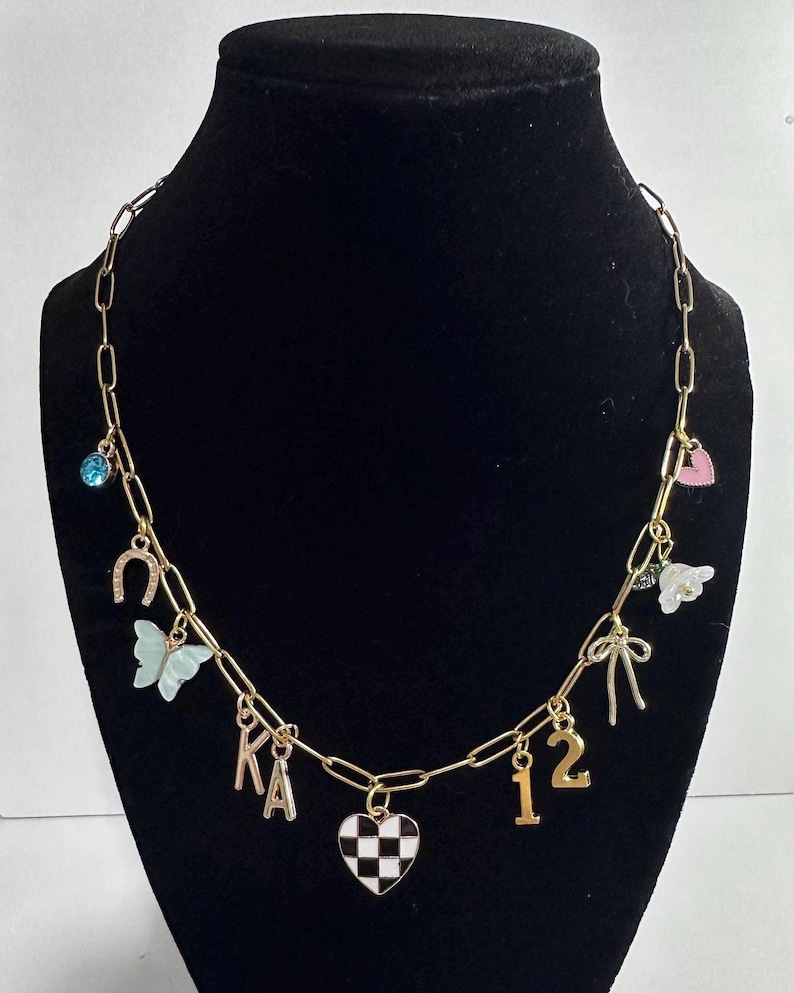 May include: A gold-toned chain necklace featuring various charms including a blue rhinestone, a horseshoe, a light blue butterfly, the letters KA, a black and white checkered heart, the number 12, a gold bow, a white flower, and a pink heart.