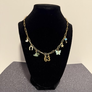 May include: A gold-toned chain necklace featuring multiple charms including a white heart, a horseshoe, a green flower, the number 63, a light blue butterfly, a small star, and a light blue key with a heart-shaped top attached to the links.