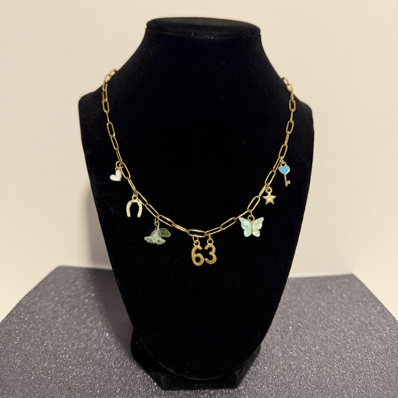May include: A gold-toned chain necklace featuring multiple charms including a white heart, a horseshoe, a green flower, the number 63, a light blue butterfly, a small star, and a light blue key with a heart-shaped top attached to the links.