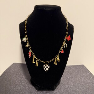 May include: A gold-toned chain necklace featuring multiple charms including a white flower, a gold star, a red lightning bolt, the letters L and H, a black and white checkered heart, the number 44, a small red heart, a horseshoe, and a red butterfly.