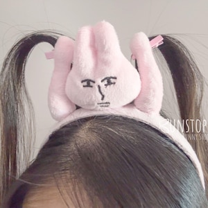 Peut inclure: Un serre-tête rose avec des oreilles de lapin et un visage de dessin animé brodé en noir. Le bandeau est porté sur la tête d'une personne avec des cheveux foncés coiffés en couettes. Le texte "UNSTOPPABLE BUNNY SHOP" est visible.