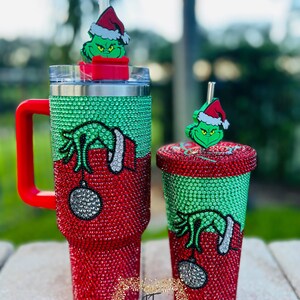 May include: Two Christmas-themed tumblers, one large with a handle and one smaller, both covered in red and green rhinestones. Each features a Grinch design and a matching Grinch topper on the straw. The larger tumbler has a red handle.