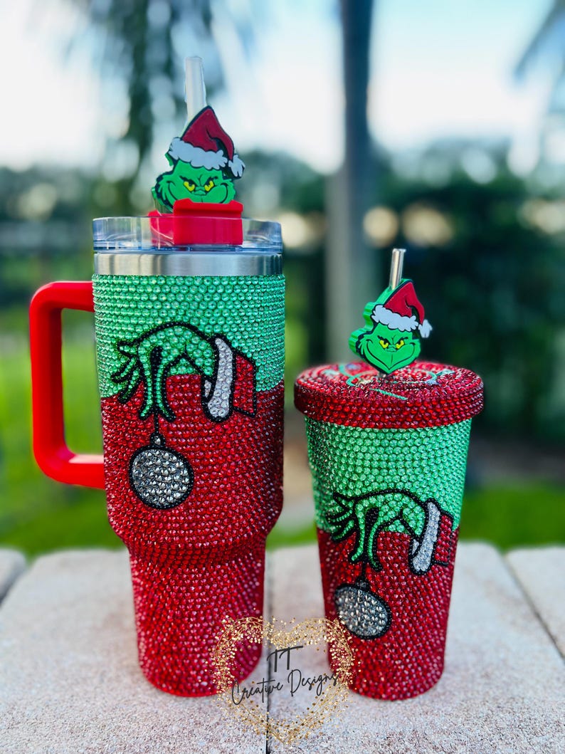 May include: Two Christmas-themed tumblers, one large with a handle and one smaller, both covered in red and green rhinestones. Each features a Grinch design and a matching Grinch topper on the straw. The larger tumbler has a red handle.