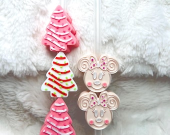 May include: Two reusable straws with festive Christmas tree and Minnie Mouse themed toppers. The Christmas tree toppers are pink, white, and green with red and white stripes. The Minnie Mouse toppers are pink and white with black bows and glitter accents.