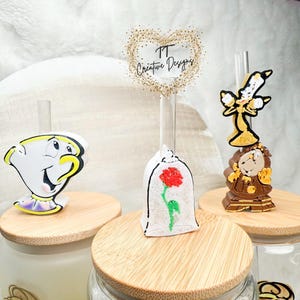 May include: Three glass drinking straws with decorative toppers featuring characters from Beauty and the Beast. The toppers are a yellow teacup with a face, a white dome with a red rose, and a gold clock with a brown face.