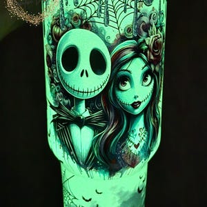 May include: Glow-in-the-dark tumbler featuring Jack Skellington and Sally from The Nightmare Before Christmas. The design includes cobwebs, bats, and a heart tattoo. The tumbler has a teal and black color scheme.