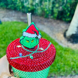 May include: A bedazzled tumbler with a Grinch-themed design. The top is red with a green Grinch character and a Santa hat. The bottom half is green. A silver straw is inserted into the tumbler.