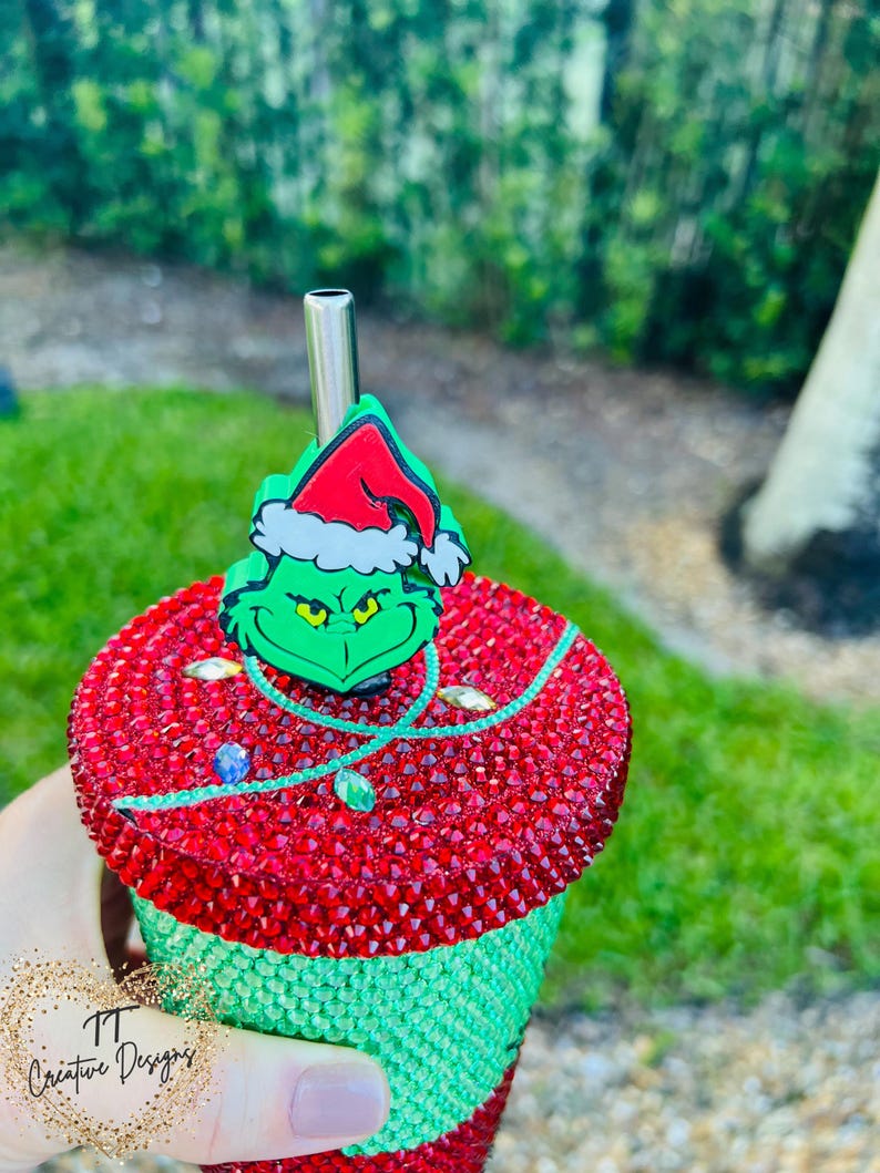 May include: A bedazzled tumbler with a Grinch-themed design. The top is red with a green Grinch character and a Santa hat. The bottom half is green. A silver straw is inserted into the tumbler.