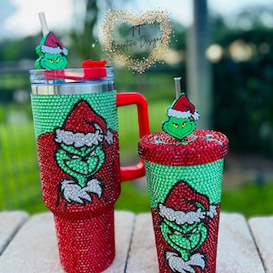 May include: Two Christmas-themed tumblers. One is a large travel mug with a red handle, and the other is a smaller cup. Both are covered in red and green rhinestones and feature a Grinch design. The tumblers have matching Grinch-themed toppers.