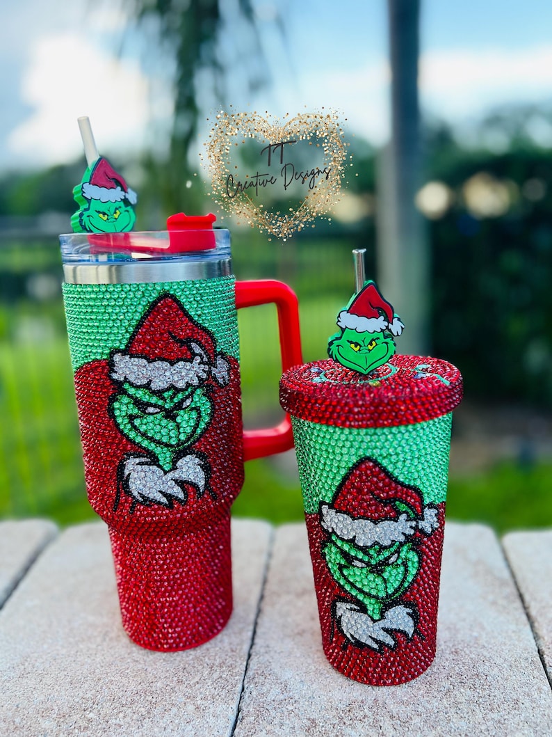 May include: Two Christmas-themed tumblers. One is a large travel mug with a red handle, and the other is a smaller cup. Both are covered in red and green rhinestones and feature a Grinch design. The tumblers have matching Grinch-themed toppers.