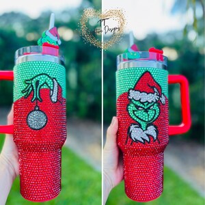 May include: A red and green tumbler with a Grinch design, covered in sparkling rhinestones. The tumbler has a red handle and a matching lid with a straw. The Grinch is wearing a Santa hat. The tumbler has the text "TT Two Designs".