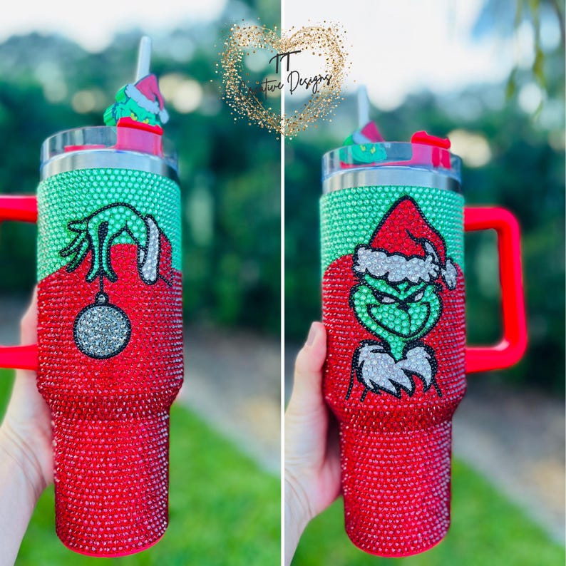 May include: A red and green tumbler with a Grinch design, covered in sparkling rhinestones. The tumbler has a red handle and a matching lid with a straw. The Grinch is wearing a Santa hat. The tumbler has the text "TT Two Designs".