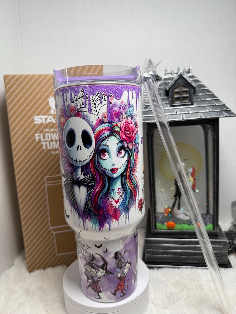 May include: A white tumbler with a purple lid and straw features Jack Skellington and Sally from The Nightmare Before Christmas. The design includes a watercolor style image of the characters, spiderwebs, and bats. The tumbler has the text "STA FLOW TUM".