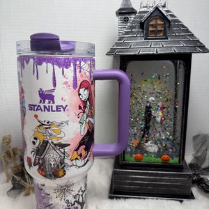 May include: A white Stanley tumbler with a purple handle and lid, featuring a colorful design of characters from The Nightmare Before Christmas. The design includes Sally, Jack Skellington, and other spooky elements. A decorative Halloween-themed snow globe is in the background.