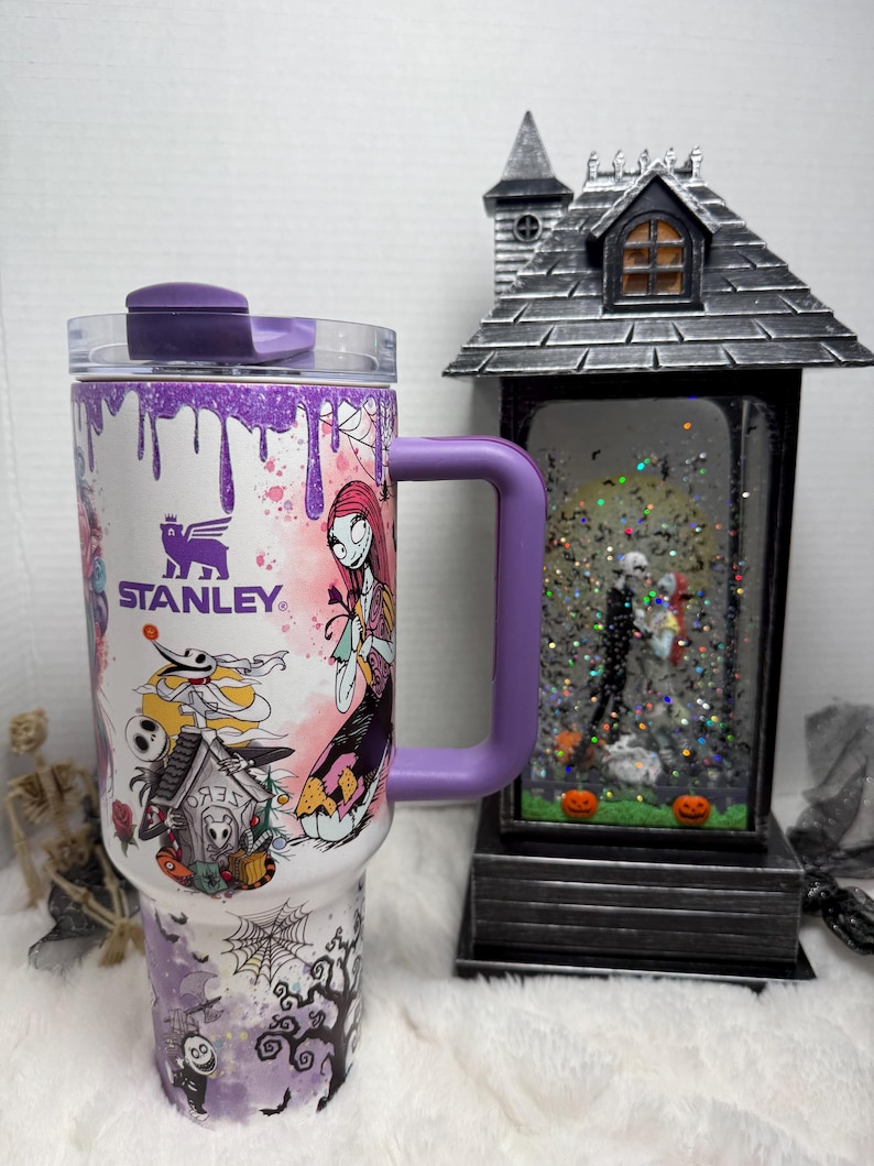 May include: A white Stanley tumbler with a purple handle and lid, featuring a colorful design of characters from The Nightmare Before Christmas. The design includes Sally, Jack Skellington, and other spooky elements. A decorative Halloween-themed snow globe is in the background.