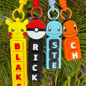 May include: Four colorful keychains featuring popular cartoon characters. The keychains include a yellow Pikachu, a red and white Pokeball, a blue Squirtle, and an orange Charmander. Each keychain has a matching lanyard and a name displayed.