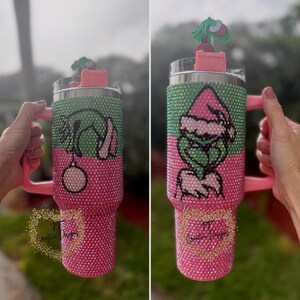 May include: A bedazzled tumbler with a pink handle and base, and a green and pink upper section. The tumbler features a Grinch design with a Santa hat and a Christmas ornament. A small Grinch figure sits on the lid.