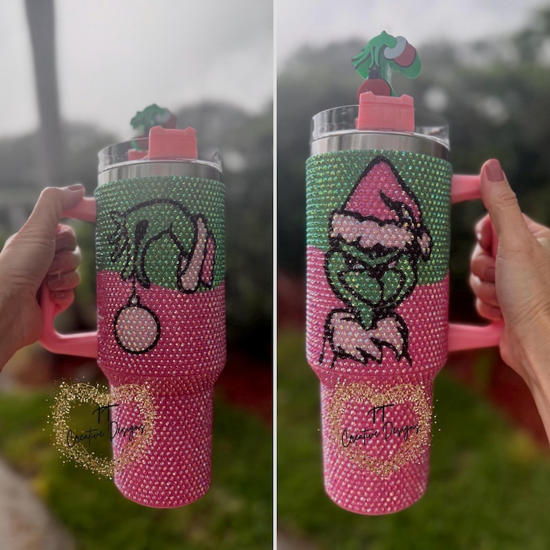 May include: A bedazzled tumbler with a pink handle and base, and a green and pink upper section. The tumbler features a Grinch design with a Santa hat and a Christmas ornament. A small Grinch figure sits on the lid.