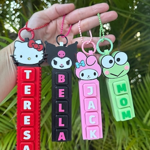 May include: Four character keychains featuring cartoon characters. The keychains are red, black, pink, and green. The red keychain says "TERESA", the black keychain says "BELLA", the pink keychain says "JACK", and the green keychain says "MOM".