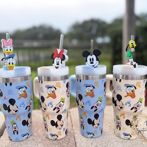 May include: Four stainless steel tumblers with character-themed toppers. The tumblers feature Mickey Mouse, Donald Duck, and Pluto designs on a light blue and cream background. Each tumbler has a handle and a straw.
