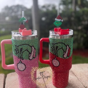 May include: Two bedazzled tumblers with Grinch-themed designs. One is pink and green, the other red and green. Each has a Grinch charm on the straw and a matching handle. The tumblers are decorated with rhinestones.