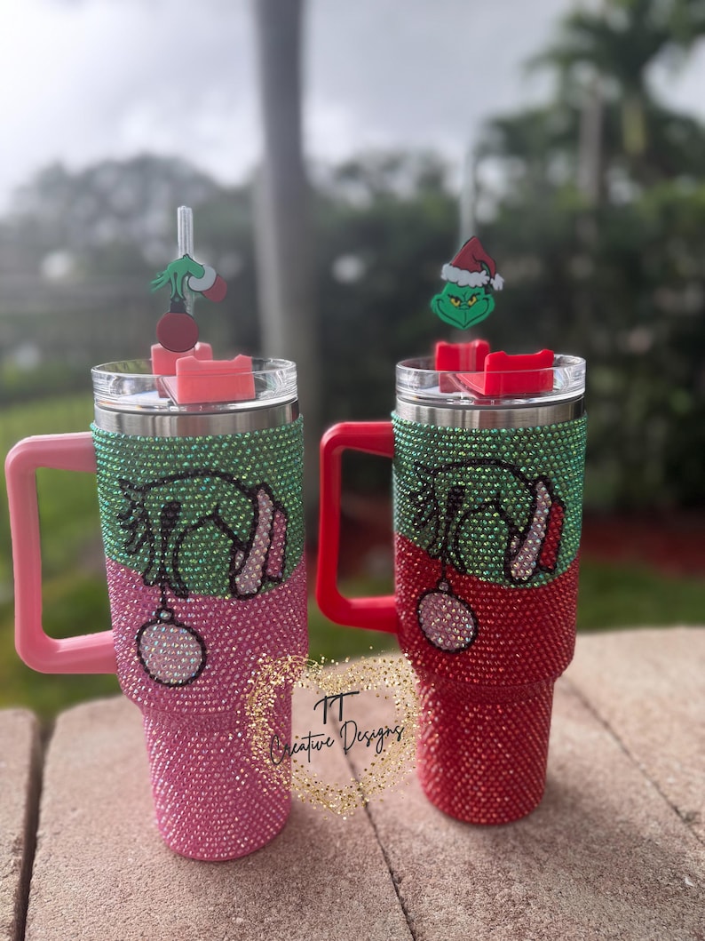 May include: Two bedazzled tumblers with Grinch-themed designs. One is pink and green, the other red and green. Each has a Grinch charm on the straw and a matching handle. The tumblers are decorated with rhinestones.