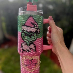 May include: A bedazzled tumbler with a pink and green Grinch design. The tumbler has a pink handle and a matching lid with a Grinch-themed topper. The bottom of the tumbler has the text "TT Creative Designs" in gold glitter.