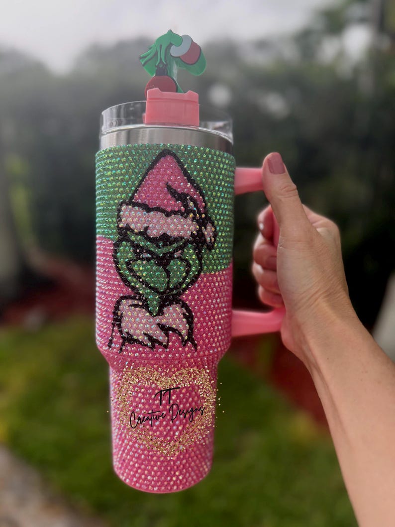 May include: A bedazzled tumbler with a pink and green Grinch design. The tumbler has a pink handle and a matching lid with a Grinch-themed topper. The bottom of the tumbler has the text "TT Creative Designs" in gold glitter.