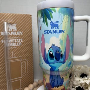 May include: A Stanley Flowstate tumbler featuring a colorful design of Stitch from Lilo & Stitch. The tumbler has a white handle and lid, and the design includes a jungle scene with butterflies. The Stanley logo is visible on the tumbler.