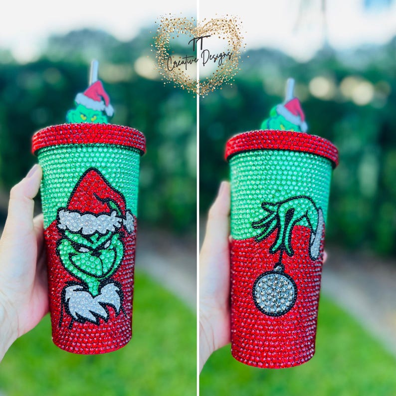 May include: A green and red bedazzled tumbler featuring the Grinch. The tumbler has a red top and bottom, with a green center covered in rhinestones. The Grinch is depicted on the front, and his hand holding an ornament on the back.