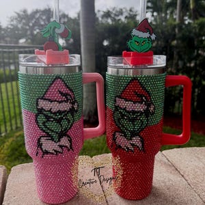 May include: Two bedazzled tumblers, one pink and one red, each featuring a Grinch design. The tumblers have green and silver accents, with a Grinch figure on the lid. The pink tumbler has a pink handle, and the red tumbler has a red handle.