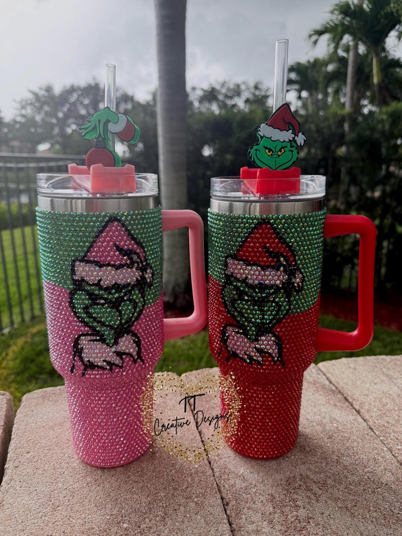 May include: Two bedazzled tumblers, one pink and one red, each featuring a Grinch design. The tumblers have green and silver accents, with a Grinch figure on the lid. The pink tumbler has a pink handle, and the red tumbler has a red handle.