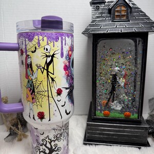 May include: A purple and white tumbler featuring characters from The Nightmare Before Christmas. The tumbler has a handle and a clear lid. A decorative Halloween-themed snow globe is also in the image.