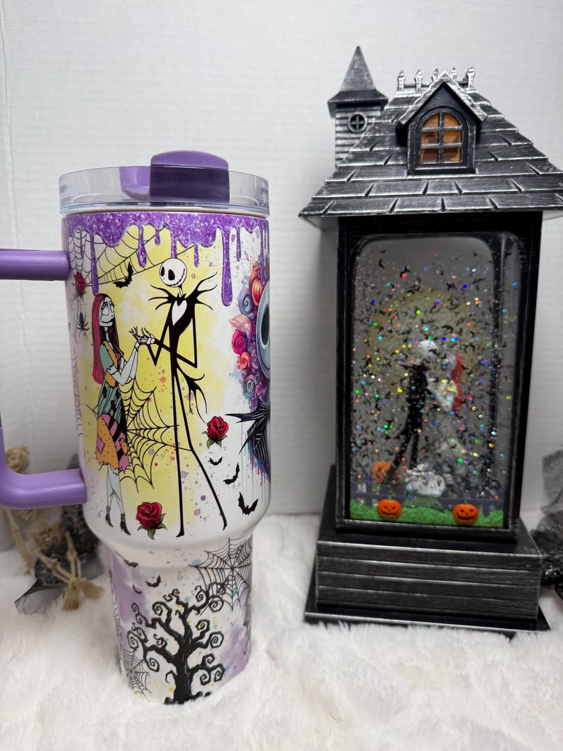 May include: A purple and white tumbler featuring characters from The Nightmare Before Christmas. The tumbler has a handle and a clear lid. A decorative Halloween-themed snow globe is also in the image.