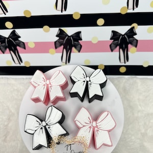 May include: Four handmade hair bows in pink, white, and black. The bows are arranged on a white surface with a gold glitter background. The text "Creative Designs" is visible in the bottom right corner of the image.