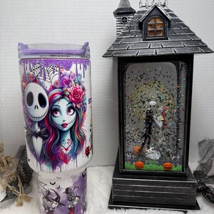 May include: A white tumbler with a purple lid featuring Jack Skellington and Sally from The Nightmare Before Christmas. A black and gray Halloween-themed snow globe with Jack and Sally inside, and two orange pumpkins. The tumbler has a purple and white design.