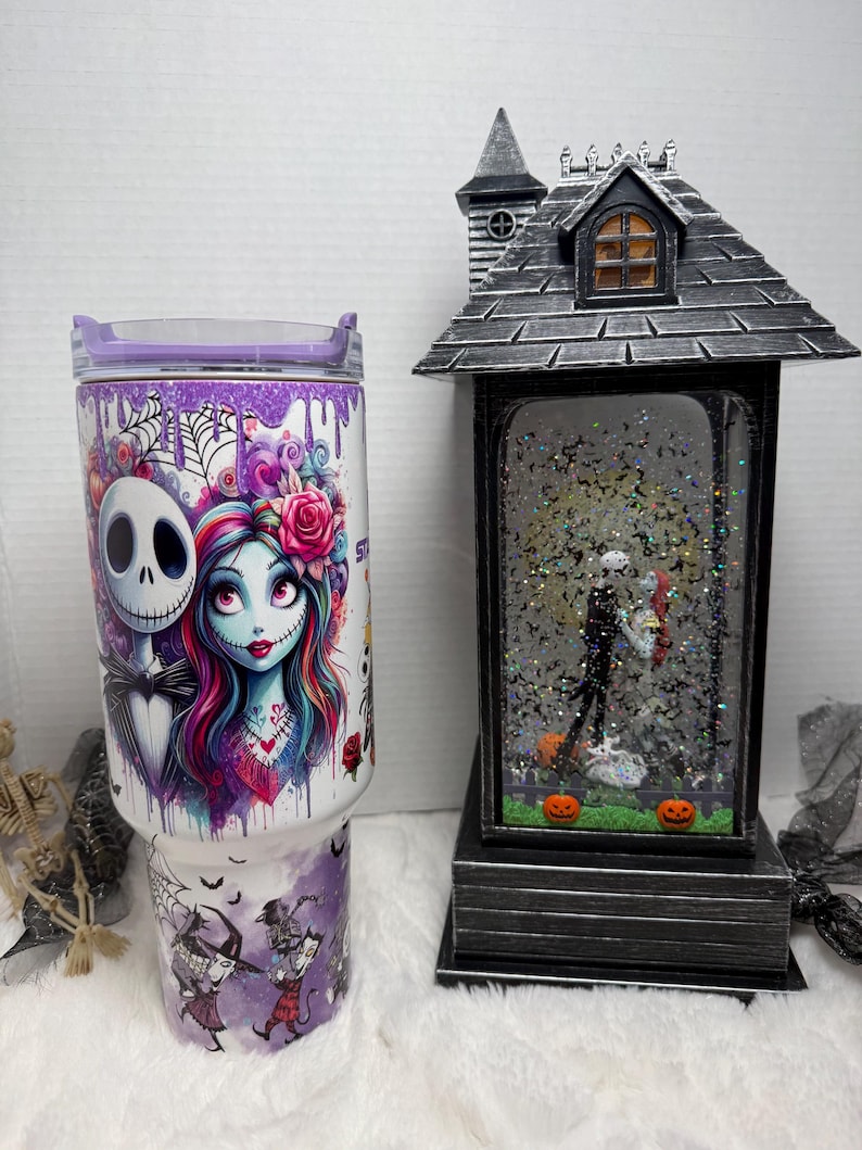 May include: A white tumbler with a purple lid featuring Jack Skellington and Sally from The Nightmare Before Christmas. A black and gray Halloween-themed snow globe with Jack and Sally inside, and two orange pumpkins. The tumbler has a purple and white design.