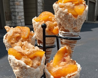 May include: Four individual peach cobbler desserts in waffle cones, displayed on a black metal stand. The cones are topped with a white chocolate drizzle and a crumb topping.