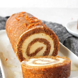 May include: A carrot cake roll with cream cheese filling, cut to show the spiral design. The cake is a warm brown color, topped with orange carrot shavings and a white glaze, presented on a white rectangular plate.