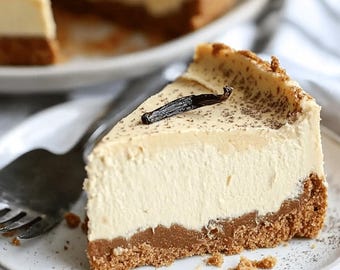 Pode incluir: Uma fatia de cheesecake num prato branco. A sobremesa tem uma base de bolacha, um recheio cremoso branco e um topo castanho claro. Uma vagem de baunilha escura está em cima da fatia. Um garfo está no prato, com migalhas espalhadas.