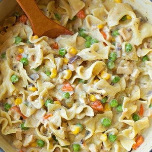 Può includere: Primo piano di una pentola blu piena di un cremoso stufato di pollo e noodles. Lo stufato è guarnito con piselli, mais, carote e funghi.