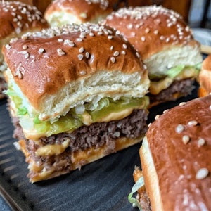 May include: Close-up of mini burgers on a black grill pan. The burgers are topped with lettuce, cheese, and a sesame seed bun.